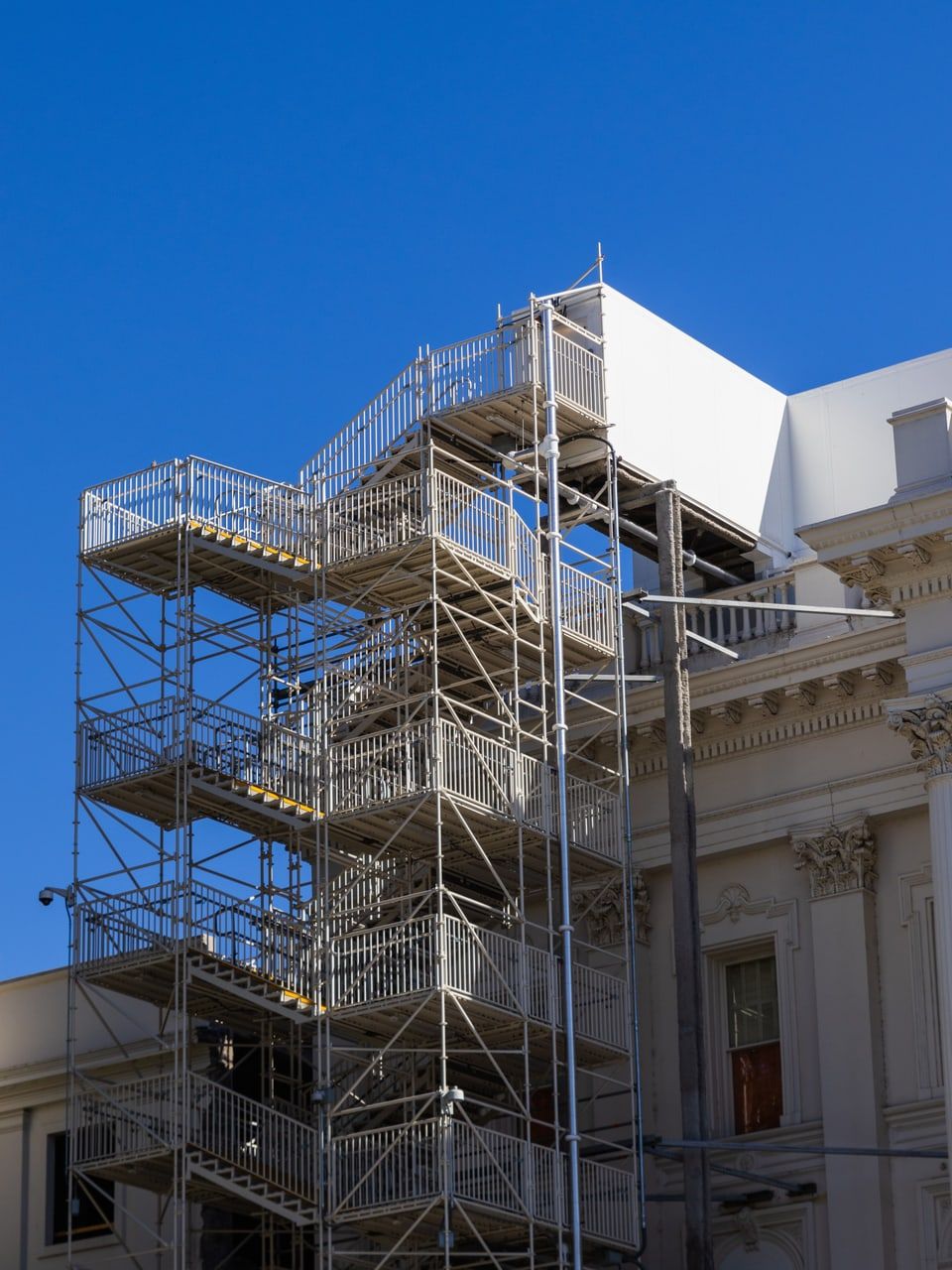 Gerüstturm mit mehreren Treppenläufen und Podesten an der Ecke eines hellen Wohngebäudes, vor blauem Himmel