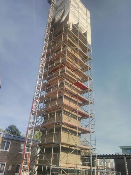 Schmaler Gerüstturm ohne Plane, an jeder Lage ein Holzbelag mit rotem Bordbrett, links ein Leiteraufstieg